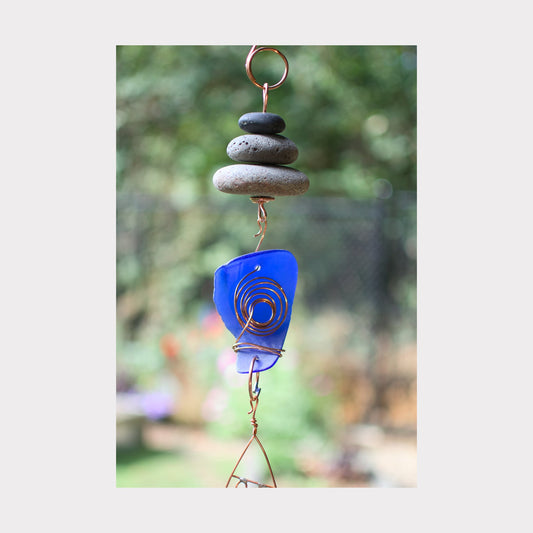 Decorative wind chime with blue glass piece and stones against a blurred natural background