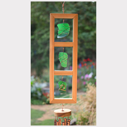 Decorative wind chime with green glass leaves and wooden frame against a garden background