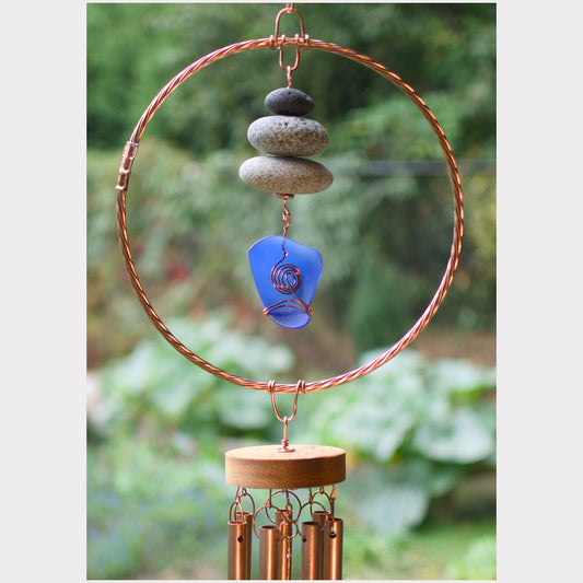 A handmade wind chime hanging ornament with a large copper-colored circular hoop, stacked stones, a blue guitar pick, and a wooden base holding copper wind chimes, set against a blurred green garden background.