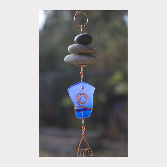 Decorative hanging with blue glass piece, stones, and copper elements against a blurred natural background