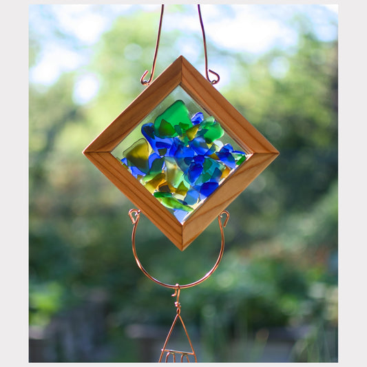 Decorative glass suncatcher with colored glass pieces in a wooden frame, hanging against a blurred green outdoor background.