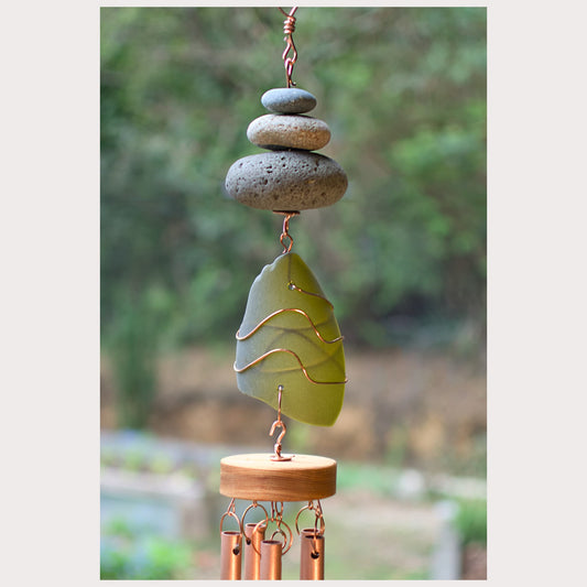Decorative wind chime with stacked stones and a green leaf-shaped ornament against a blurred natural background.