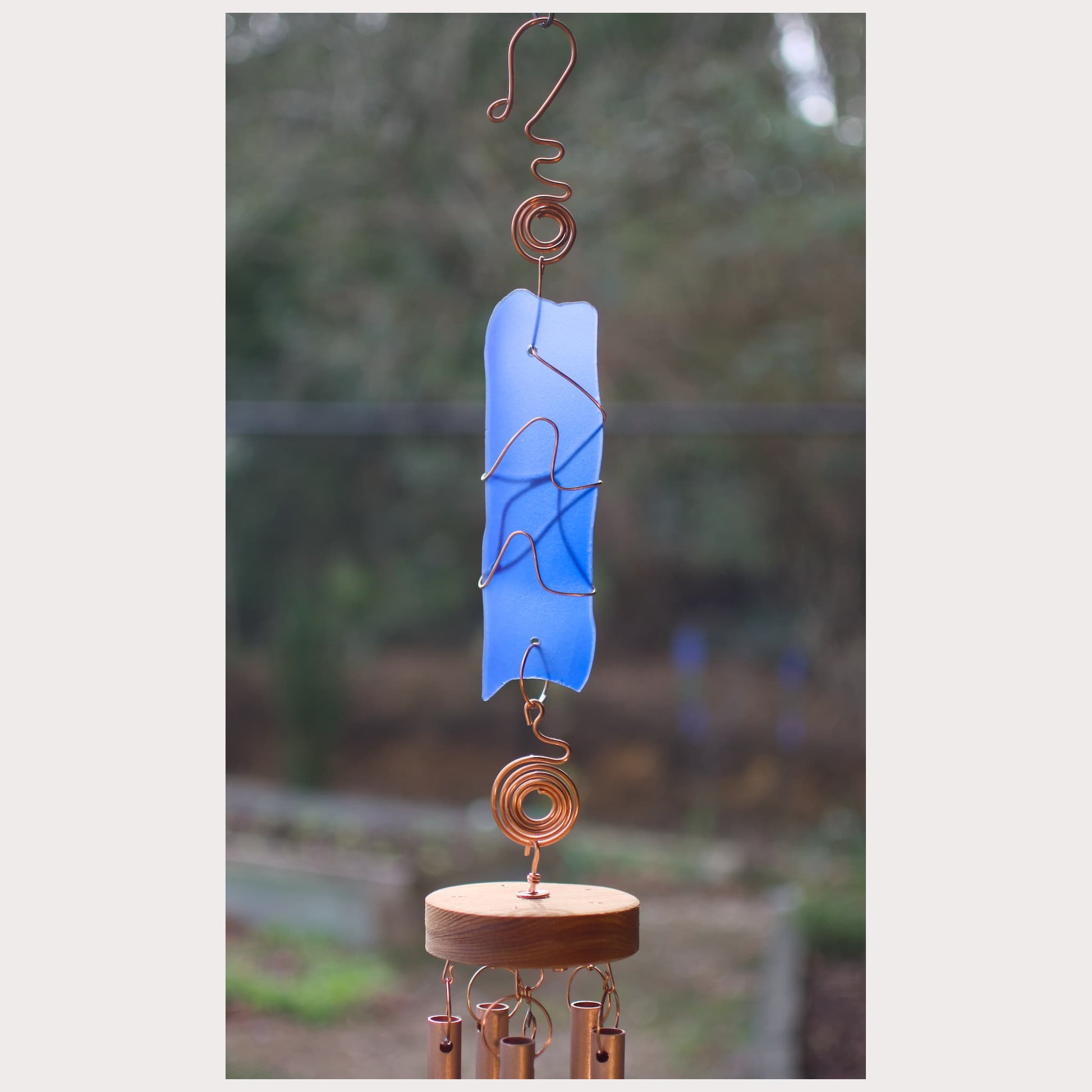 Decorative wind chime with blue glass panel and copper elements against a blurred natural background