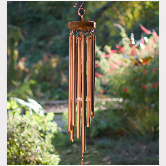 A copper wind chime hanging outdoors in a garden setting with blurred green foliage and plants in the background.
