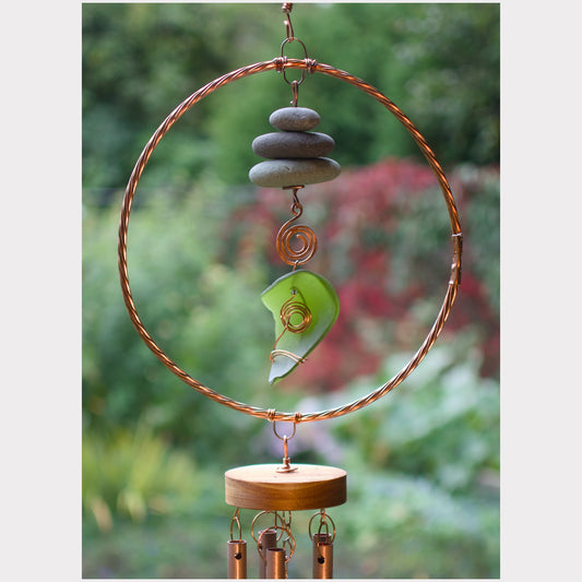 A handmade wind chime hanging outdoors against a blurred green garden background. The chime features a large copper wire hoop, a stack of gray stones, a green glass piece, and a round wooden bottom plate with hanging copper tubes.
