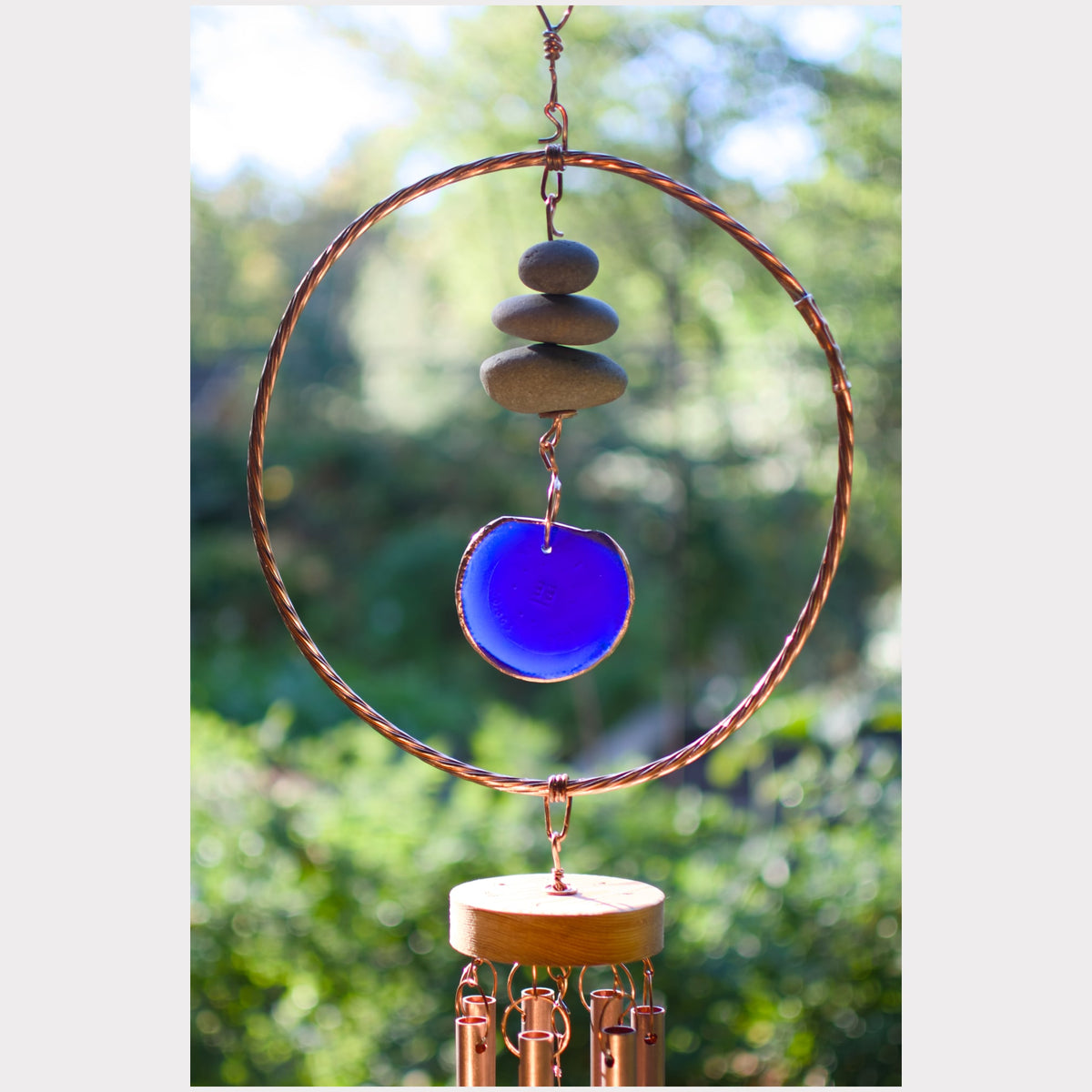 A copper wind chime with a large circular hoop, stacked stones, a vibrant blue glass disc, and copper wind chime tubes, set against a blurred green garden background.