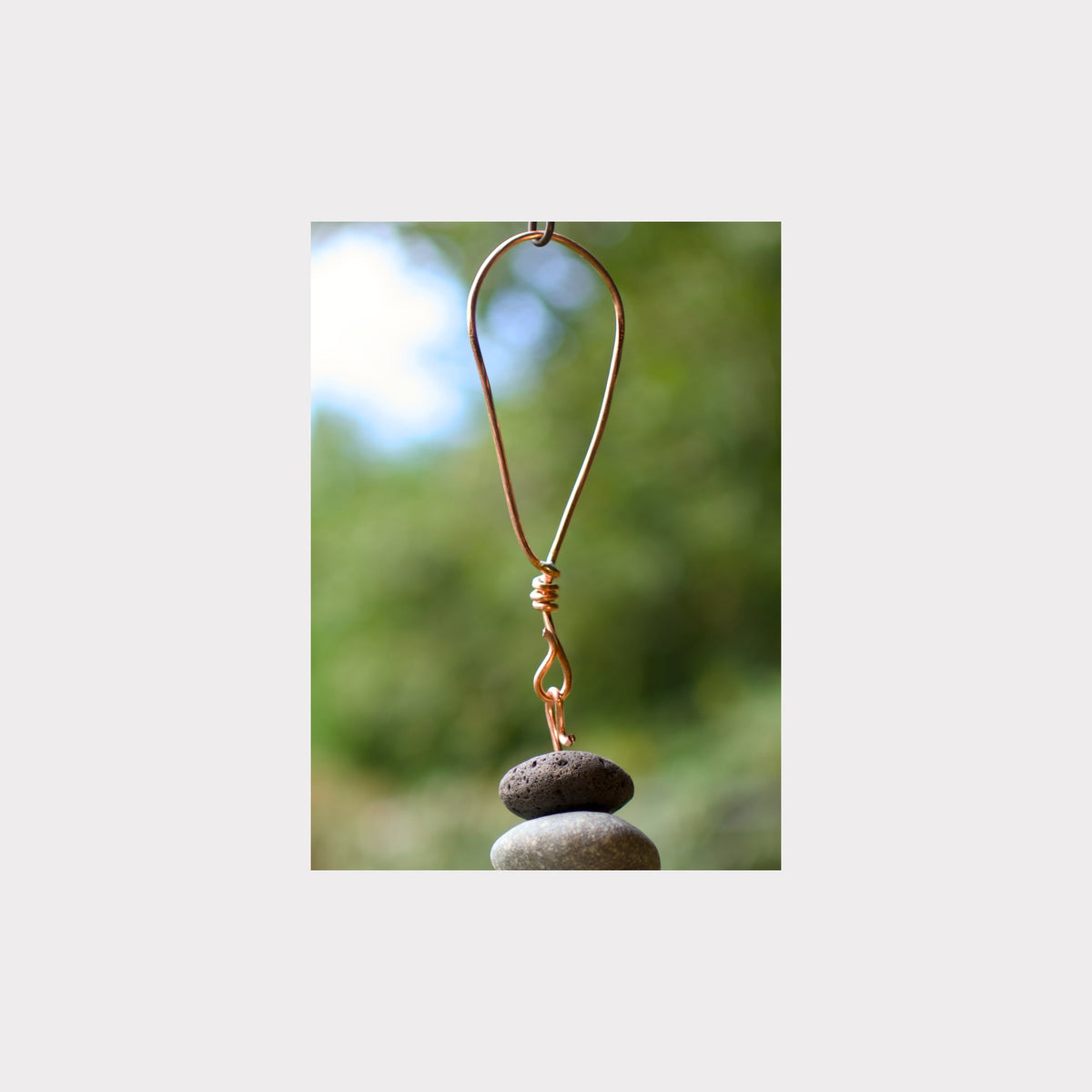 A rustic metal wind chime featuring stacked dark gray stones suspended by a copper-colored wire loop, shown against a blurred green outdoor background.