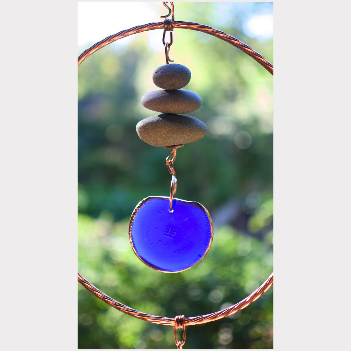 A handmade bohemian wind chime featuring three stacked gray stones, a round cobalt blue glass disc, and twisted copper wire hoops against a blurred green outdoor background.