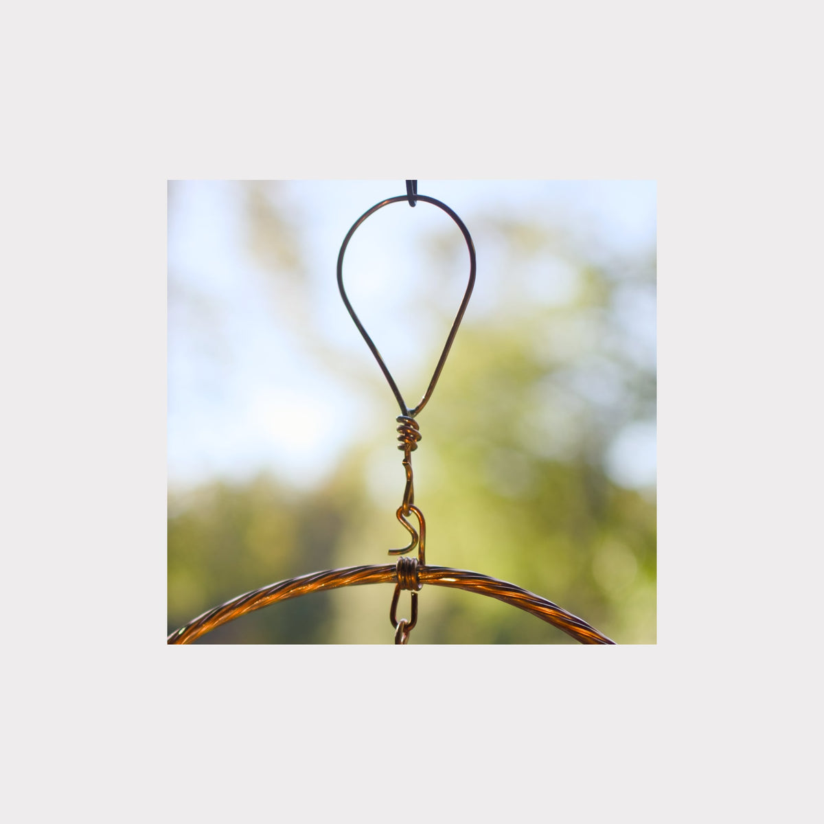 A close-up of a metal hanging loop with a twisted wire connector and a curved metal bar, shown against a soft blurred outdoor background.