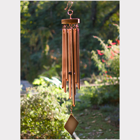 A copper wind chime with multiple hanging tubes and a diamond-shaped pendant, shown outdoors against a blurred garden background.