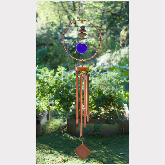 A copper wind chime hanging outdoors in a garden, featuring a circular hoop with stacked stones and a blue glass disc, alongside multiple copper-colored metal tubes and a diamond-shaped end piece.