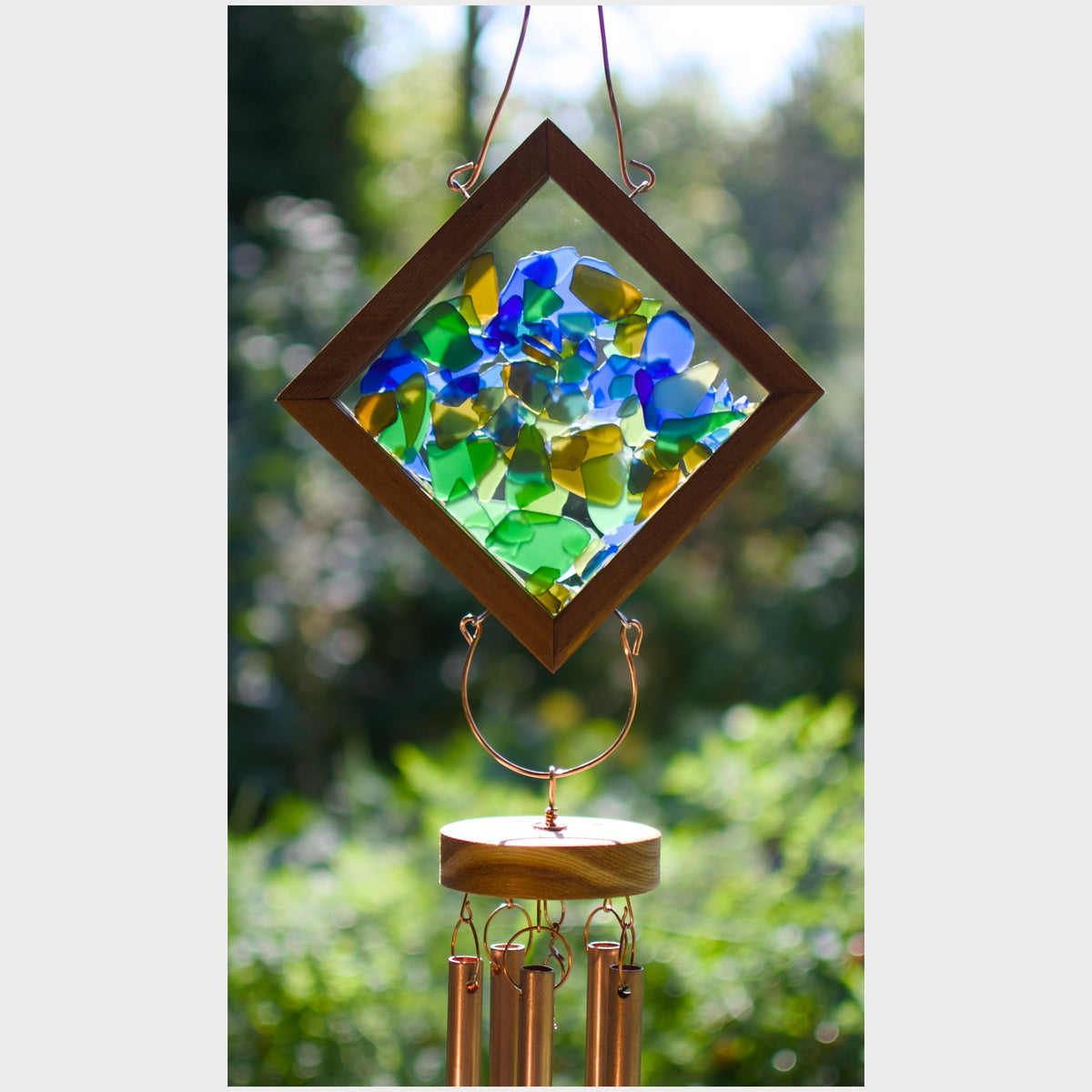 A diamond-shaped hanging wind chime with a wooden frame and colorful stained-glass style panels in blue, green, and yellow, suspended over copper-colored wind chime tubes against a blurred outdoor garden background.