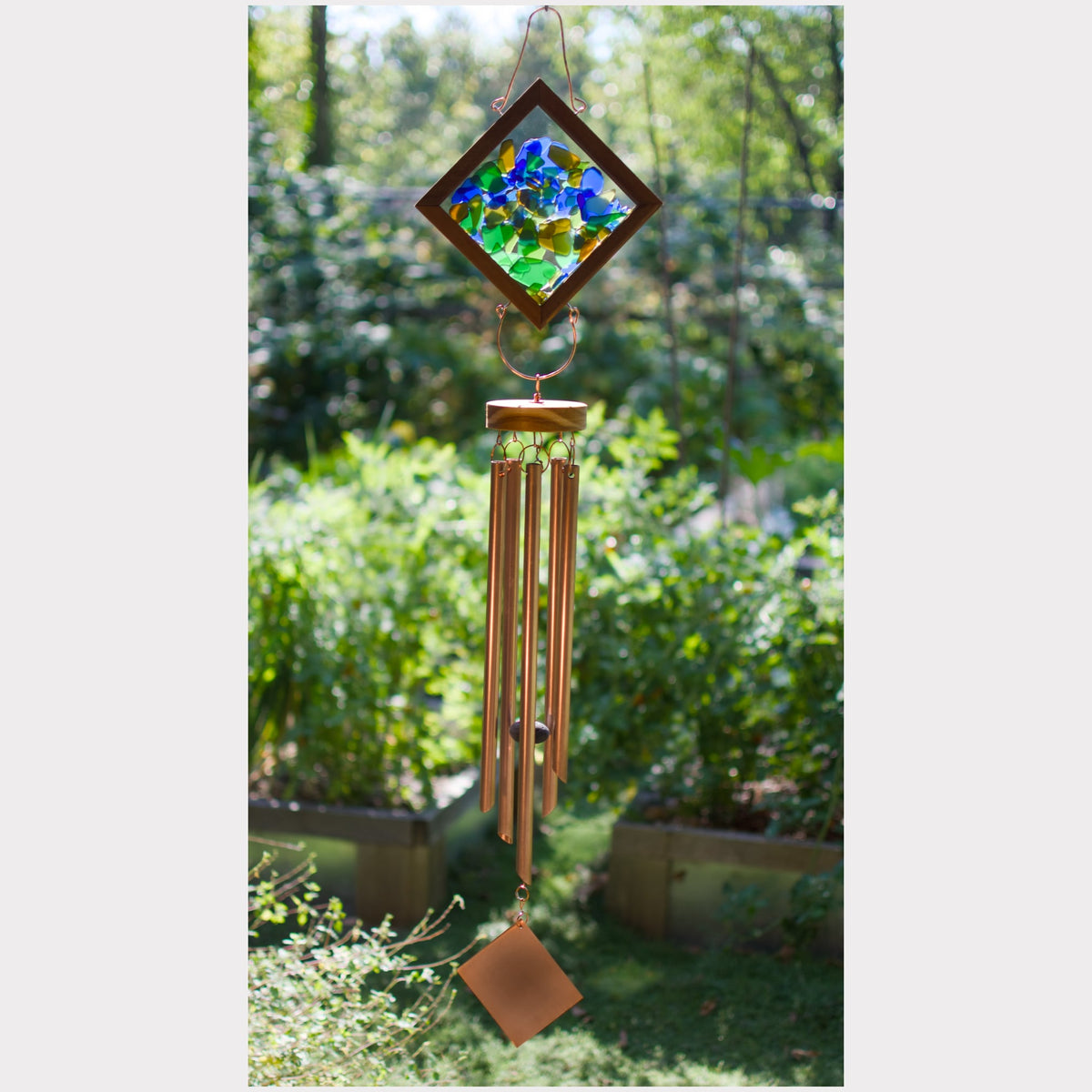 A hanging wind chime with a diamond-shaped stained-glass top featuring blue, green, and yellow pieces, copper-toned metal tubes, and a small diamond-shaped copper pendant, photographed outdoors against a blurred garden background.